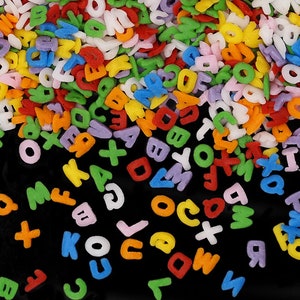 Puede incluir: Una dispersión de coloridos sprinkles con letras del alfabeto sobre un fondo negro. Los sprinkles son de varios colores, incluyendo rojo, naranja, amarillo, verde, azul, morado y blanco.