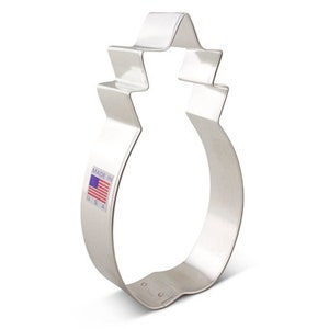 Könnte beinhalten: Ein silberner Keksausstecher in Form einer Ananas. Der Ausstecher hat einen gezackten Rand und einen kleinen Aufkleber mit der amerikanischen Flagge, auf dem "Made in U.S.A." steht.