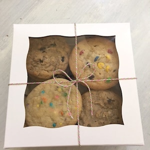 Op de afbeelding: Een witte doos met een doorzichtig venster, vastgebonden met touw, met vier verschillende koekjes. De koekjes zijn chocoladekoekjes, koekjes met sprankelende suiker, M&M-koekjes en een gewoon koekje.