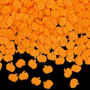 May include: A close-up shot of orange pumpkin-shaped sprinkles scattered on a black surface. The sprinkles are small, with a smooth texture and a bright, uniform color. These edible decorations are perfect for Halloween-themed treats.