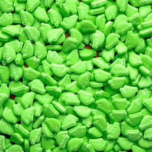 Puede incluir: Una gran cantidad de caramelos de color verde brillante con forma de rodajas de sandía. Los caramelos están esparcidos sobre una superficie.