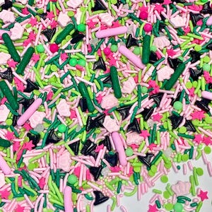 Op de afbeelding: Een mix van groene, roze en zwarte sprinkles in verschillende vormen, waaronder sterren, stokjes en kronen.