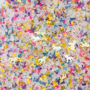 May include: A close-up of a mixture of colorful sprinkles, including white, pink, blue, purple, and gold. The sprinkles are in the shape of stars, squares, and unicorns.