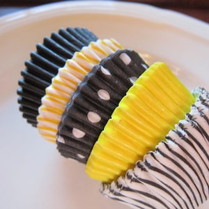 Könnte beinhalten: Ein Stapel Cupcake-Förmchen in schwarz, weiß und gelben Mustern. Die Förmchen sind aus Papier und haben eine geriffelte Form.