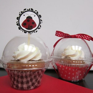 May include: Two clear plastic cupcake containers with red and white polka dot cupcake liners. One cupcake has a ladybug topper and the other has a red ribbon.