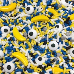 May include: A close-up of colorful sprinkles, including yellow banana shapes, blue and white sprinkles, and white frosting with black eyes.