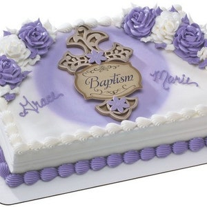 Puede incluir: Un pastel cuadrado blanco con glaseado morado y blanco. El pastel está decorado con una cruz dorada con la palabra "Bautismo" en ella. El pastel también tiene los nombres "Grace" y "Marie" escritos en él.