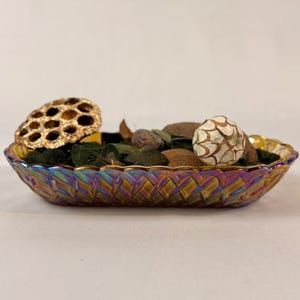 May include: An oval-shaped iridescent glass dish with a woven pattern, filled with dried botanicals. The dish contains a variety of items, including a dried lotus pod, small stones, and a decorative ball. The dish has a gold rim.