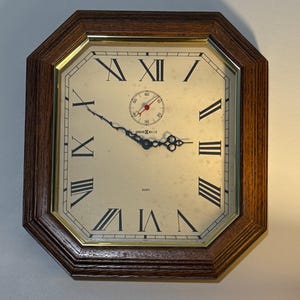 May include: Octagonal quartz clock with a dark brown wooden frame and a cream-colored face. The clock features black Roman numerals, a small second dial, and the brand name "Howard Miller". The hands are black and ornate.