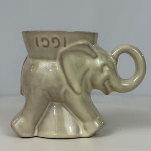 May include: A ceramic mug in the shape of an elephant, with a light beige glaze. The mug features the year "1991" embossed on the rim. The handle is a loop, and the elephant's trunk forms the spout.