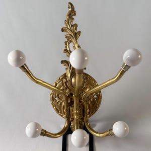 May include: A wall-mounted coat rack featuring a gold-toned metal base with ornate, swirling filigree detailing. The rack has five extending arms, each tipped with a smooth, round white ceramic knob, providing multiple points for hanging items.