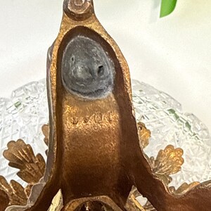 May include: A bronze-toned metal stand featuring a textured, leaf-like design and a circular base. The metal surface displays a weathered patina with visible markings and a dark, circular inset at the top. A clear, faceted glass element is visible.
