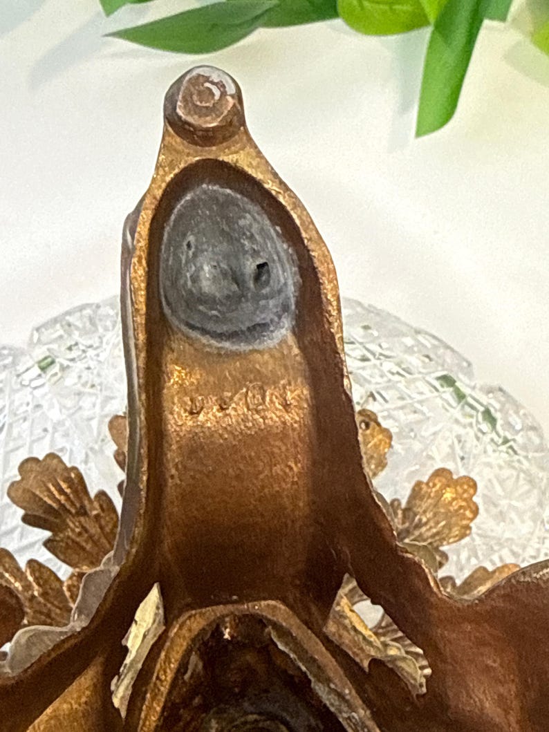 May include: A bronze-toned metal stand featuring a textured, leaf-like design and a circular base. The metal surface displays a weathered patina with visible markings and a dark, circular inset at the top. A clear, faceted glass element is visible.