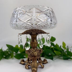 May include: A clear cut glass bowl with a geometric diamond pattern sits atop a decorative antique bronze-toned metal pedestal base. The base features ornate leaf-like detailing and is adorned with hanging clear teardrop-shaped glass crystals.