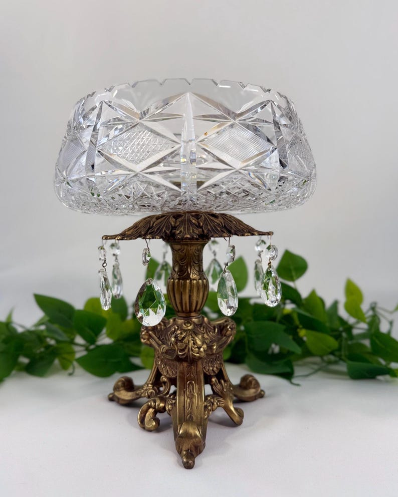 May include: A clear cut glass bowl with a geometric diamond pattern sits atop a decorative antique bronze-toned metal pedestal base. The base features ornate leaf-like detailing and is adorned with hanging clear teardrop-shaped glass crystals.