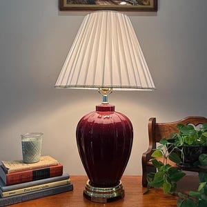May include: A table lamp with a glossy, deep red, vertically ribbed ceramic base and a polished gold-tone metal pedestal. The lamp features a white, pleated, conical fabric lampshade with a delicate beaded trim along the bottom edge.