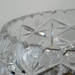 May include: A clear cut glass bowl featuring a complex geometric pattern of intersecting lines, starburst motifs, and diamond-shaped facets. The rim of the bowl has a notched, serrated edge, and the glass surface reflects light through its deep cuts.