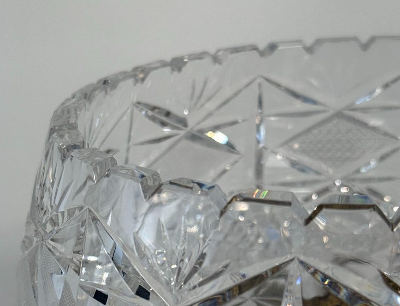 May include: A clear cut glass bowl featuring a complex geometric pattern of intersecting lines, starburst motifs, and diamond-shaped facets. The rim of the bowl has a notched, serrated edge, and the glass surface reflects light through its deep cuts.