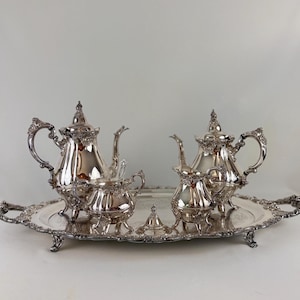 May include: A silver tea set consisting of a large teapot, a smaller teapot, a creamer, a sugar bowl with a lid, and a serving tray. Each piece features ornate, embossed floral detailing along the edges, handles, spouts, and feet.