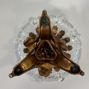 May include: A bottom view of a decorative object featuring a three-legged brass base with dark circular pads at each tip. The base has an ornate, leaf-like metal design surrounding a central opening, positioned beneath a clear, diamond-cut glass bowl.