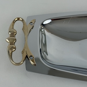 May include: A rectangular serving tray with a polished silver-tone finish and a reflective surface. The tray features a decorative gold-tone metal handle attached to the side with two visible screws, showcasing a curved, symmetrical design.