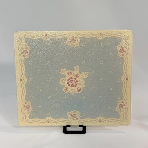 May include: A rectangular glass cutting board featuring a textured surface with a light blue center and a cream-colored border. The design includes a central rose motif and smaller floral accents along the edges, framed by a wavy line pattern.