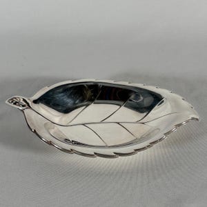May include: A polished silver-toned metal dish shaped like a leaf. The dish features a smooth, reflective surface with embossed vein details running through the center and extending toward the serrated edges, mimicking a natural foliage form.