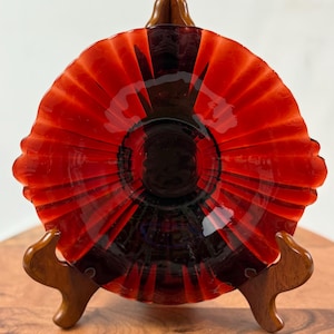 May include: A vibrant red glass bowl with a fluted design, featuring a dark center. The bowl is displayed on a wooden stand. The bowl's rim has a scalloped edge. The bowl is likely for decorative or serving purposes.