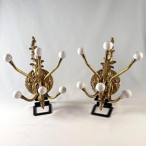 May include: A pair of wall-mounted coat racks featuring ornate gold-toned metal backplates with intricate scrollwork. Each rack has five curved arms extending outward, with each arm tipped by a smooth, round white ceramic ball finial.