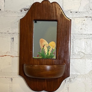 May include: A wooden wall-mounted shelf with a mirror and a small bowl-shaped shelf. The mirror features a painting of mushrooms and grass. The wood has a dark brown stain and is mounted on a white brick wall.