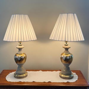 May include: A pair of table lamps featuring white pleated fabric shades and urn-shaped bases. The bases are finished in a combination of matte gray and metallic gold, with a hand-painted white floral pattern on the gold section and a textured base.