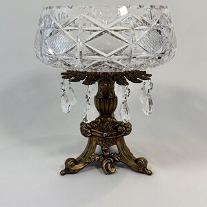 May include: A clear cut glass bowl with a diamond and cross-hatch pattern sits atop a decorative bronze-toned metal pedestal base. The base features ornate scrollwork and is adorned with several dangling clear crystal teardrop prisms.
