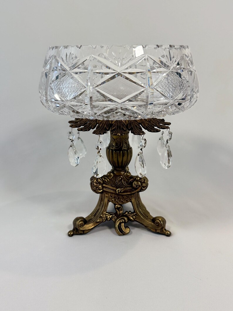 May include: A clear cut glass bowl with a diamond and cross-hatch pattern sits atop a decorative bronze-toned metal pedestal base. The base features ornate scrollwork and is adorned with several dangling clear crystal teardrop prisms.