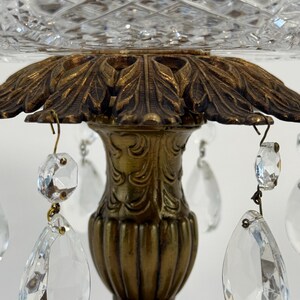 May include: A decorative centerpiece featuring a clear cut-glass bowl with a diamond pattern, supported by an ornate brass-toned metal pedestal base. The base is adorned with hanging clear crystal teardrop prisms attached by metal hooks.