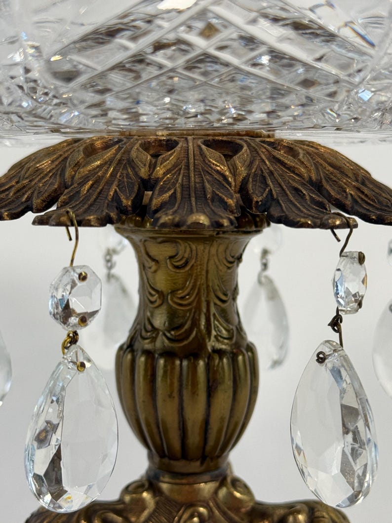 May include: A decorative centerpiece featuring a clear cut-glass bowl with a diamond pattern, supported by an ornate brass-toned metal pedestal base. The base is adorned with hanging clear crystal teardrop prisms attached by metal hooks.