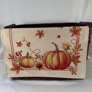 May include: A rectangular woven table runner featuring a beige background with a printed design of two orange pumpkins, sunflowers, red berries, and autumn leaves in shades of red, orange, and yellow, framed by thin vertical orange stripes.