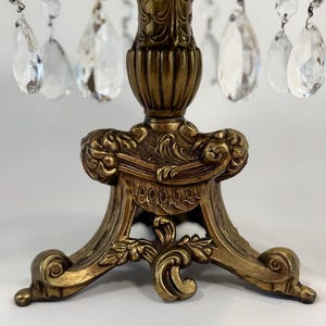 May include: A decorative stand featuring an antique gold-toned metal base with ornate, scrolled relief detailing. The stand is adorned with multiple clear, teardrop-shaped glass crystals suspended from the top rim by small metal rings.