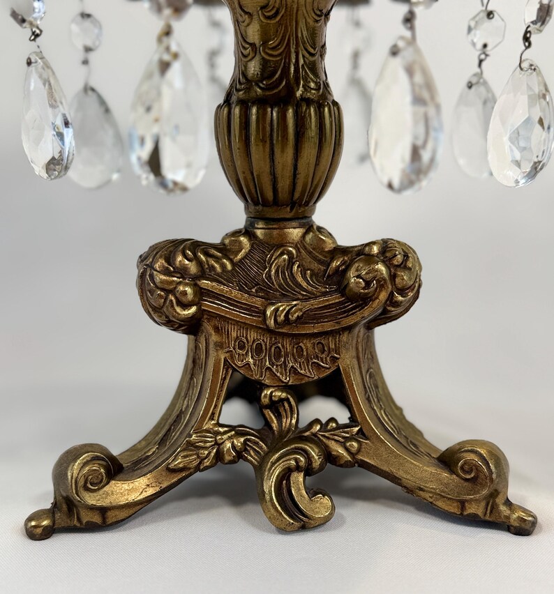 May include: A decorative stand featuring an antique gold-toned metal base with ornate, scrolled relief detailing. The stand is adorned with multiple clear, teardrop-shaped glass crystals suspended from the top rim by small metal rings.