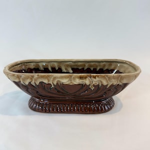 May include: A brown ceramic planter with a rectangular shape and a decorative floral pattern. The planter has a raised base with vertical lines and a rim with a light brown glaze.