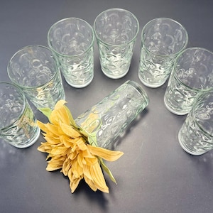 May include: Eight clear drinking glasses with a textured pattern are arranged in a semi-circle. A yellow flower with green leaves rests near the center. The glasses are on a dark gray surface.