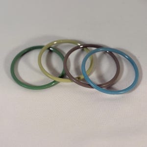 May include: A set of four thin, circular bangle bracelets in varying colors. The collection includes one dark green, one light olive green, one dark brown, and one bright blue bangle, each featuring a smooth, solid, and glossy finish.