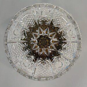 May include: A circular clear crystal bowl featuring an intricate cut glass pattern of diamonds and stars. The center of the bowl is adorned with a decorative antique brass-toned metal insert featuring a detailed floral and leaf motif design.