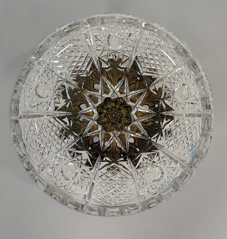 May include: A circular clear crystal bowl featuring an intricate cut glass pattern of diamonds and stars. The center of the bowl is adorned with a decorative antique brass-toned metal insert featuring a detailed floral and leaf motif design.