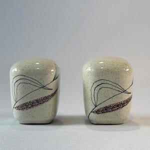 May include: A pair of off-white ceramic salt and pepper shakers with a speckled finish. Each shaker features a black line drawing of a curved leaf and a textured, dark brown stippled pattern along the base, creating a cohesive set of kitchenware.