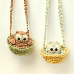 May include: Two crocheted owl ornaments, each sitting in a small bowl-shaped basket suspended by a braided cord. One owl is brown with a cream face in a sage green basket, and the other is light gray with a white face in a mustard yellow basket.