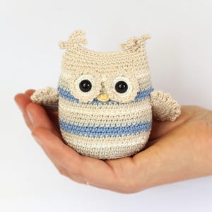 Puede incluir: Una pequeña figura de búho de ganchillo en beige, blanco y azul. El búho tiene grandes ojos negros y un pequeño pico amarillo. Las alas y los mechones de las orejas están detallados. El búho se sostiene en una mano.