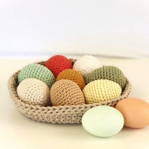 May include: A woven basket filled with colorful, crocheted eggs in shades of red, green, yellow, brown, and white. Two real eggs, one light green and one tan, sit beside the basket. The eggs are a decorative Easter theme.