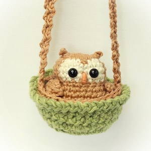May include: A small, crocheted brown and cream-colored owl with large black eyes and an orange beak, sitting inside a light green crocheted basket. The basket is suspended by two braided tan yarn straps that extend upward from the sides.