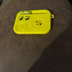 May include: Bright yellow, square-shaped case with rounded edges. The case features a circular design in the center, two small circular buttons, and a rectangular bar at the top. A small loop is on the side. The case is likely for electronic devices.