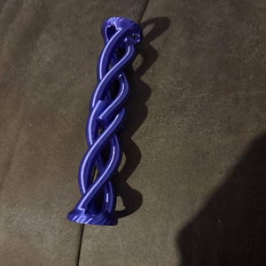 Op de afbeelding: Een paarse decoratieve flesstopper met een gedraaid, touwachtig ontwerp. De stopper heeft een uitlopende basis en een lus aan de bovenkant. Het ontwerp is een continu, verweven patroon, waardoor een unieke visuele textuur ontstaat.